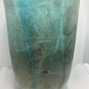 Op de afbeelding: Een hoge, cilindervormige vaas met een afgeronde basis. De buitenkant heeft een gevlekte turquoise en blauwgroene glazuur met donkere strepen, wat een textuur creëert. De bovenste rand is helderder turquoise.