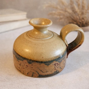 May include: A handmade ceramic jug with a rounded body, a small handle, and a flared opening. The pottery features a tan and brown colour scheme with a dark blue band near the base. The piece is likely for decorative or functional use.