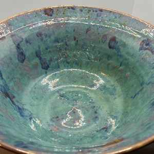 Peut inclure: Un bol en céramique fait à la main avec une finition brillante et texturée. Le bol présente un mélange de teintes turquoise, bleues et brunes, avec un bord doré. L'intérieur a un aspect moucheté, suggérant une création artisanale unique.