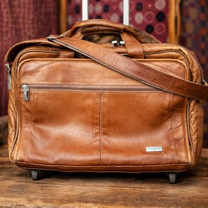 Könnte beinhalten: Eine braune Leder-Rollaktentasche mit Teleskopgriff und Schulterriemen. Die Tasche hat eine Fronttasche mit Reißverschluss und ein kleines Metallschild. Die Aktentasche steht auf einer Holzoberfläche.