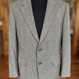 Peut inclure: Un blazer en tweed gris avec une fermeture à deux boutons, présenté sur un mannequin noir. Le blazer a deux poches avant et un revers cranté. Le tissu présente un motif moucheté.