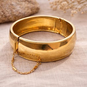 以下が含まれることがあります： 金色のヒンジ付きバングルブレスレット。ブレスレットは、滑らかで光沢のある表面を持つ幅広のバンドです。小さなゴールドチェーンが留め金に取り付けられています。ジュエリーは明るい色の生地に展示されています。