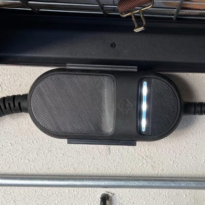 Puede incluir: Un cargador de vehículo eléctrico negro con una superficie texturizada y una luz blanca brillante. El cargador está montado en una pared blanca y tiene un cable negro.