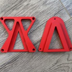 May include: Two bright red, geometric shapes with a triangular design and rounded edges. Each shape has small holes at the corners. The objects are on a grey wooden surface.