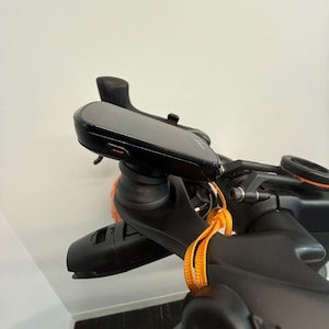 May include: A black cycling computer mounted on a bicycle handlebar stem. The device features a dark screen and a small red indicator light on the side. A bright orange lanyard is looped around the black handlebar stem assembly.