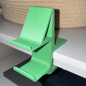 May include: A bright green shelf clip, designed to hold items on a shelf. The clip has a unique shape with two curved arms to secure items. The clip is made of a solid material and is painted in a vibrant green color.