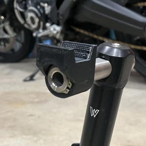 Könnte beinhalten: Schwarzer Motorradständer mit silbernem Metallakzent und schwarzer Halterung. Der Ständer hat ein 'W'-Logo an der Seite. Der Hintergrund zeigt Teile eines Motorrads.