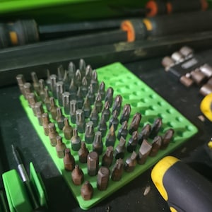 Peut inclure: Un ensemble de différents embouts de tournevis dans un support vert, entouré de tournevis et d'autres outils. Les embouts sont argentés et marron, et les tournevis ont des manches noirs et jaunes. Les outils sont disposés sur une surface sombre.