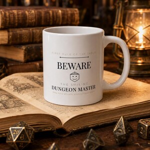 Może przedstawiać: Biały ceramiczny kubek z czarnym napisem FIRST RULE OF THE TABLE BEWARE THE SMILING DUNGEON MASTER oraz małą grafiką uśmiechniętej dwudziestościennej kości. Kubek posiada zakrzywione ucho oraz gładki, cylindryczny korpus o prostej formie.