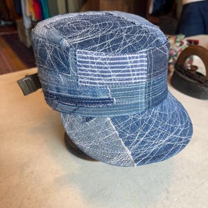 Op de afbeelding: Een pet in militaire stijl gemaakt van verschillende tinten blauwe denimstof. De hoed heeft een patchworkontwerp met zichtbare witte stikpatronen, een platte bovenkant en een korte klep. Aan de zijkant is een klein olijfgroen stoffen lipje bevestigd.
