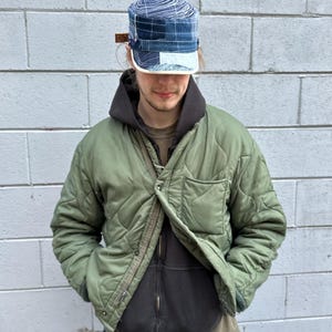 Puede incluir: Chaqueta acolchada verde oliva sobre una sudadera con capucha negra. La persona lleva una gorra estampada en azul y blanco con un borde blanco. El fondo es una pared de ladrillos gris.