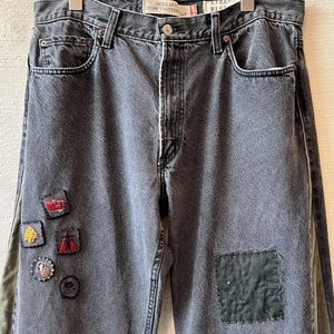 May include: A pair of charcoal grey denim jeans featuring a regular fit, a button fly, and multiple decorative patches. The front includes small embroidered patches and larger rectangular fabric patches in dark green and light blue tones.