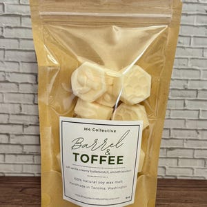 Könnte beinhalten: Ein klarer Plastikbeutel mit sechseckigen, cremefarbenen Wachsschmelzen. Das Etikett trägt die Aufschrift "Barrel & Toffee" mit den Worten "soft vanilla, creamy butterscotch, smooth bourbon" darunter. Der Beutel ist versiegelt und vor einer weißen Backsteinmauer.