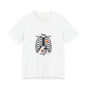Peut inclure: Un t-shirt blanc avec un motif noir représentant une cage thoracique humaine. À l'intérieur de la cage thoracique se trouve un petit squelette tenant une citrouille orange. Le design inclut un petit cœur orange, une chauve-souris et des étoiles.