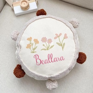 May include: A round, off-white canvas pillow featuring a printed design of three small floral stems in yellow and pink. The name Beallara is printed in pink script below the flowers. The pillow has a textured border and six small pom-poms.
