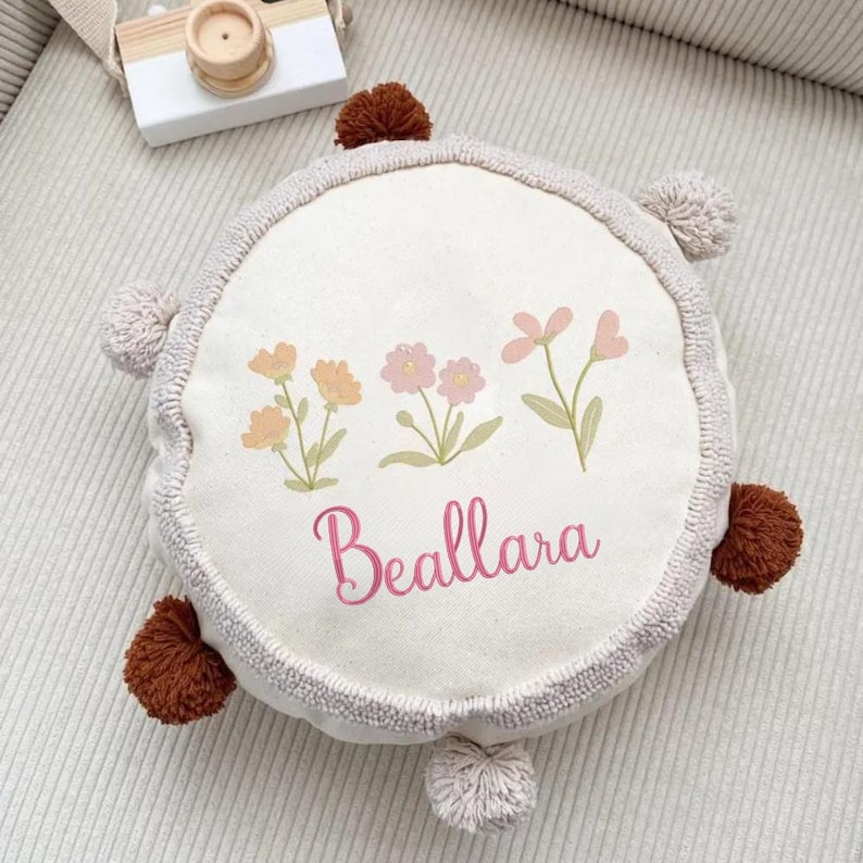 May include: A round, off-white canvas pillow featuring a printed design of three small floral stems in yellow and pink. The name Beallara is printed in pink script below the flowers. The pillow has a textured border and six small pom-poms.