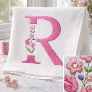 May include: White hand towel embroidered with a large pink letter "R". The letter is decorated with a floral design in pink, green, and purple. The towel is folded and displayed on a light-colored surface.