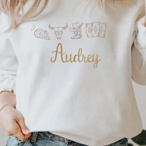 May include: A white long-sleeve sweatshirt featuring a brown line-art graphic of a cowboy hat, a cow skull, a boot, and playing cards above the name Audrey written in a cursive gold-toned font across the center of the chest area.