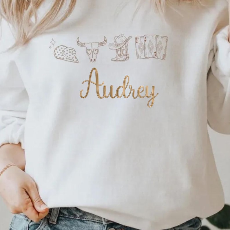 May include: A white long-sleeve sweatshirt featuring a brown line-art graphic of a cowboy hat, a cow skull, a boot, and playing cards above the name Audrey written in a cursive gold-toned font across the center of the chest area.