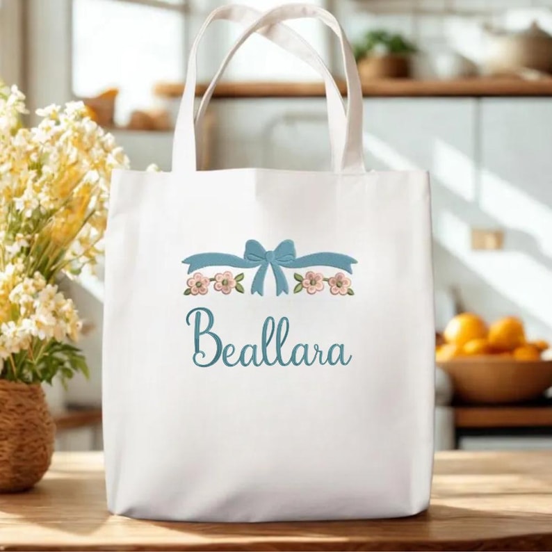 May include: A white canvas tote bag featuring a light blue embroidered bow centered above a row of small pink flowers with green leaves. Below the floral design, the name Beallara is embroidered in a cursive, light blue script font.