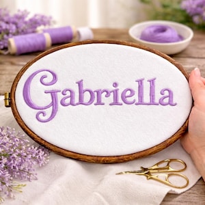May include: An oval embroidery hoop with the name "Gabriella" stitched in purple thread on white fabric. The wooden hoop is held by a hand, with gold scissors and purple thread spools nearby. The background includes sprigs of lavender.
