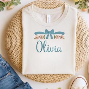 May include: A cream-colored short-sleeve t-shirt featuring an embroidered design of a light blue ribbon bow above a row of small pink flowers with green leaves. The name Olivia is embroidered in a cursive light blue font below the floral design.