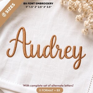 Op de afbeelding: Een witte stof met de naam "Audrey" geborduurd in een koperkleurig scriptlettertype. De tekst "BX FONT EMBROIDERY" en maatopties van 2,5 cm tot 8,9 cm staan bovenaan. De tekst "With complete set of alternate letters!" staat onderaan.