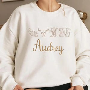 May include: A white crewneck sweatshirt featuring a brown line-art graphic of a cowboy hat, a steer skull, a boot, and three playing cards above the name Audrey written in a cursive brown script font across the center of the chest.