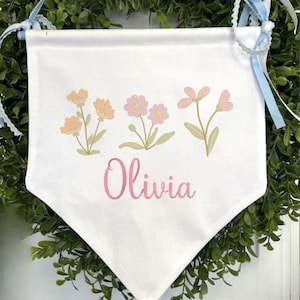 May include: A white fabric wall banner featuring a printed design of three small floral clusters in shades of peach and pink with green stems. The name Olivia is printed in a pink cursive script below the flowers. The banner has light blue ribbons.