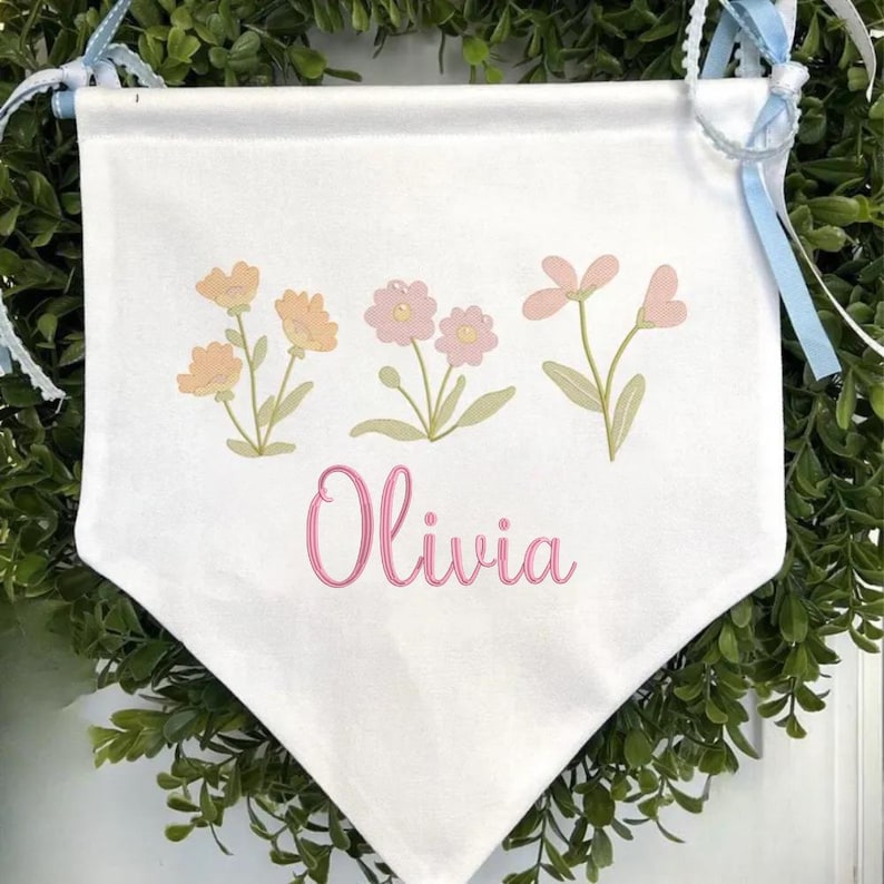 May include: A white fabric wall banner featuring a printed design of three small floral clusters in shades of peach and pink with green stems. The name Olivia is printed in a pink cursive script below the flowers. The banner has light blue ribbons.