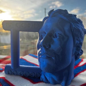 Può includere: Una scultura blu della testa di un uomo e un supporto rettangolare blu con le parole "BE GOOD" e "DO GOOD" stampate sopra. La scultura è su una superficie a strisce rosse, bianche e blu.