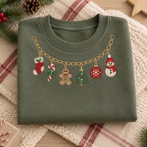 Puede incluir: Una sudadera de cuello redondo color verde oliva con un diseño bordado de una cadena dorada. La cadena presenta dijes navideños colgantes que incluyen un calcetín rojo, un bastón de caramelo, un hombre de jengibre, un árbol, una esfera y un muñeco de nieve.