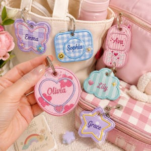 Puede incluir: Una colección de llaveros de tela bordada en varias formas, incluyendo una mariposa, un rectángulo, un corazón, una nube, una estrella y un teléfono móvil. Cada llavero presenta un nombre único: Emma, Sophia, Ava, Olivia, Lily y Grace.