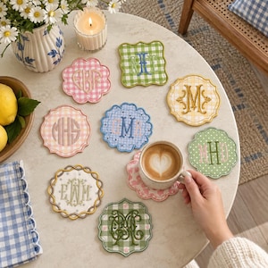 May include: A collection of nine scalloped-edge fabric coasters featuring various gingham patterns in pink, green, yellow, and blue. Each coaster is embroidered with a unique monogram in contrasting thread colors, arranged on a round table.