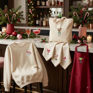 May include: A cream-colored sweatshirt with a small Santa face embroidery, a cream tote bag with a martini glass design, cream napkins with cocktail embroideries, and a dark red apron with a green martini glass embroidery, all featuring holiday themes.