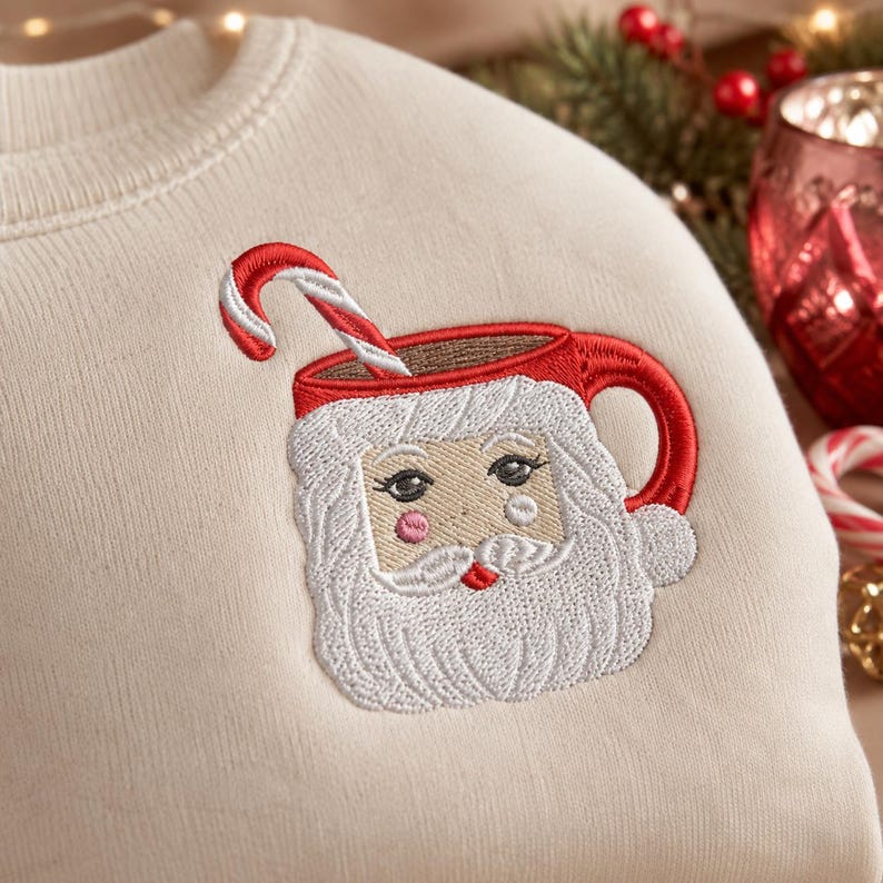 May include: A cream-colored sweatshirt featuring an embroidered design of a red mug shaped like a Santa Claus face. The Santa figure has a white beard, pink cheeks, and a red candy cane sticking out of the top of the mug. The embroidery is detailed.