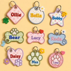 May include: Nine embroidered pet ID tags in various shapes including diamond, circle, star, bone, and paw print. Each tag features a name such as Ollie, Bella, Bobby, Bear, Lucy, Koda, Daisy, Max, or Luna, with small attached charms like flowers or hearts.
