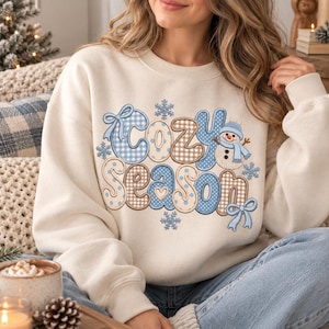 Puede incluir: Una sudadera de cuello redondo color crema con el texto "cozy season" en letras grandes estilo burbuja. Las letras están decoradas con lunares azules, patrones de cuadros marrones y lazos azul claro, junto a un pequeño muñeco de nieve y copos.