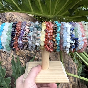 May include: A display of gemstone chip bracelets in various colors, including turquoise, green, purple, red, and white. The bracelets are arranged on a wooden stand, showcasing the variety of natural stone jewelry.