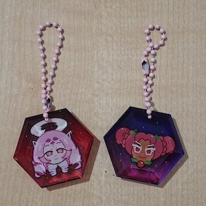 Op de afbeelding: Twee zeshoekige acrylhangers met lichtroze kogelkettingen. De linkerhanger is doorschijnend rood met een witharig personage met een halo. De rechterhanger is doorschijnend paars met een personage met rood haar in knotjes.