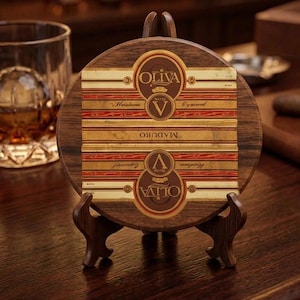 May include: A circular wooden coaster featuring a decorative Oliva Serie V Maduro cigar band design. The coaster is displayed on a small, dark brown wooden stand. The band graphic includes the text Oliva, V, and Maduro in gold and brown.