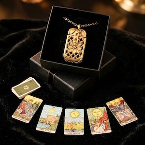 May include: A gold-toned rectangular pendant necklace with an intricate floral filigree design, presented in a black box. Below the box are five tarot cards: The Empress, The Star, The Moon, The Sun, and The Lovers, arranged in a semi-circular pattern.