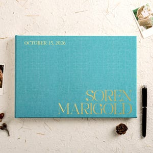 Peut inclure: Un livre turquoise avec l'inscription dorée "October 15, 2026" et "Soren Marigold". Un stylo noir et une pomme de pin sont à côté du livre. La couverture du livre est texturée.