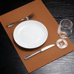 May include: A brown leather placemat with a white plate, a fork, a knife, and a wine glass. The placemat has a stitched border and is set on a dark wooden table. The plate is centered on the placemat.