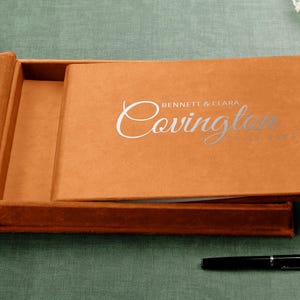 Peut inclure: Un coffret rectangulaire en velours orange brûlé contenant un album photo assorti. La couverture présente un texte en feuille d'argent indiquant "BENNETT & CLARA" au-dessus du mot "Covington" en écriture cursive et la date "10.22.2022". Un stylo noir est posé à côté.