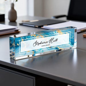 May include: A rectangular desk nameplate with a blue and gold marbled design. The nameplate has the name "Stephanie Hall" and the title "General Manager" in black script within a white frame.