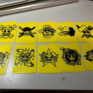May include: Ten yellow card holders with black cartoon skull and crossbones designs. Each card holder features a different skull and crossbones design, with a small tab at the top. The designs are from the One Piece anime series.