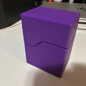 May include: A solid purple box with a geometric design. The box is rectangular with a lid that slides open. The box is sitting on a white surface.