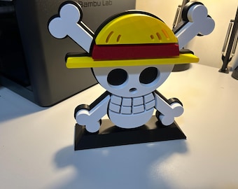 May include: A 3D-printed One Piece anime logo featuring a white skull with crossed bones, a yellow straw hat with a red band, and a black base. The design is a stylized representation of the iconic pirate flag.