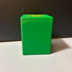 May include: A bright green, rectangular box with a lid. The top of the box features a gold-colored, stylized floral design. The box is sitting on a white surface with a black background.